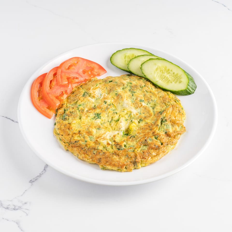 A golden omelet with herbs, served on a white plate with sliced tomatoes and cucumbers.