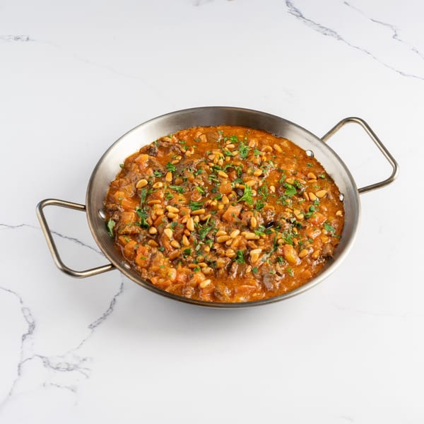 A metal pan filled with a rich, orange stew, garnished with pine nuts and fresh parsley.