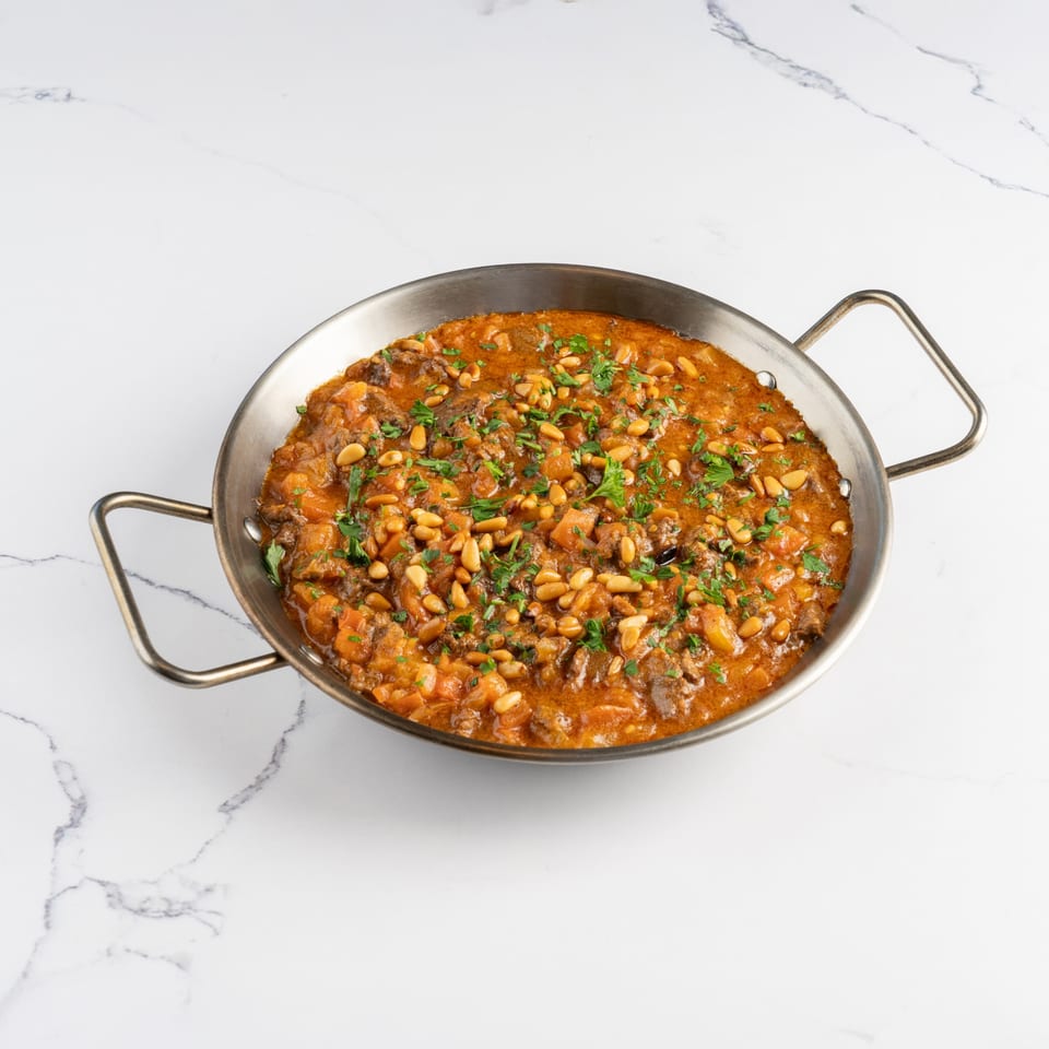A metal pan filled with a rich, orange stew, garnished with pine nuts and fresh parsley.