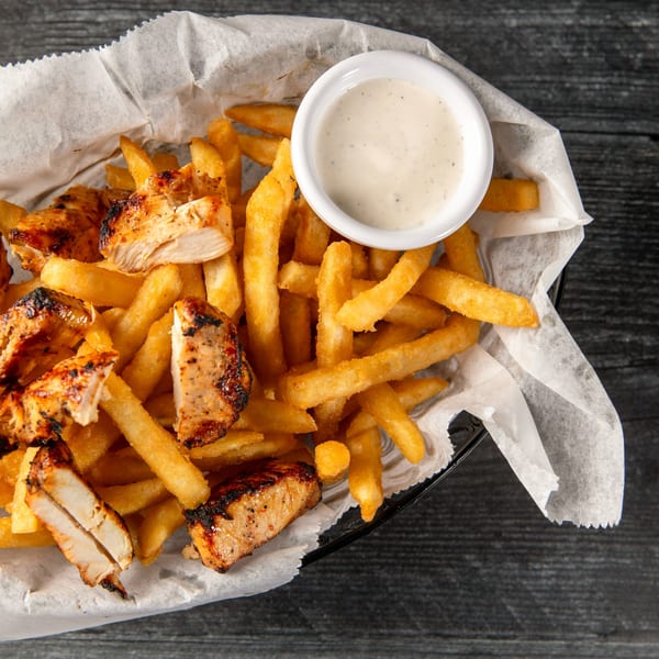 Grilled chicken pieces and golden french fries in a basket with a side of dipping sauce.