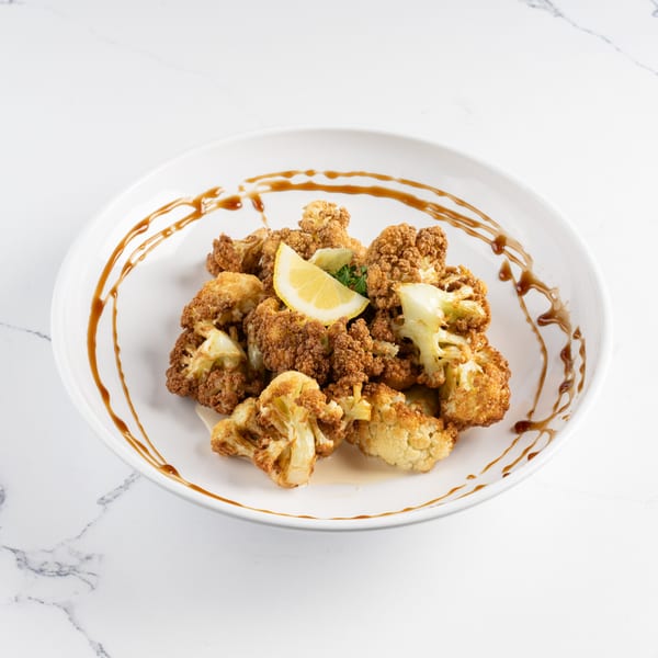 Crispy fried cauliflower florets with a lemon wedge and herb garnish, drizzled with sauce on a white plate.