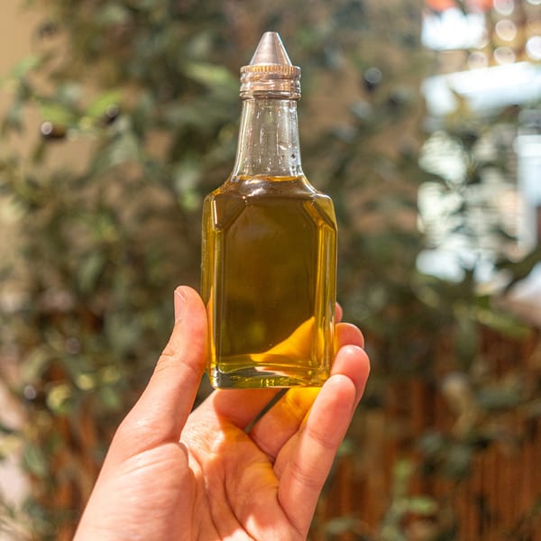 Hand holding a small glass bottle of golden olive oil with a metal pour spout.