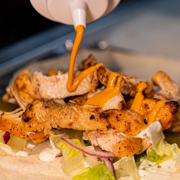 Palestinian chicken shawarma with lettuce, red onion, and white and orange sauces being drizzled on top.
