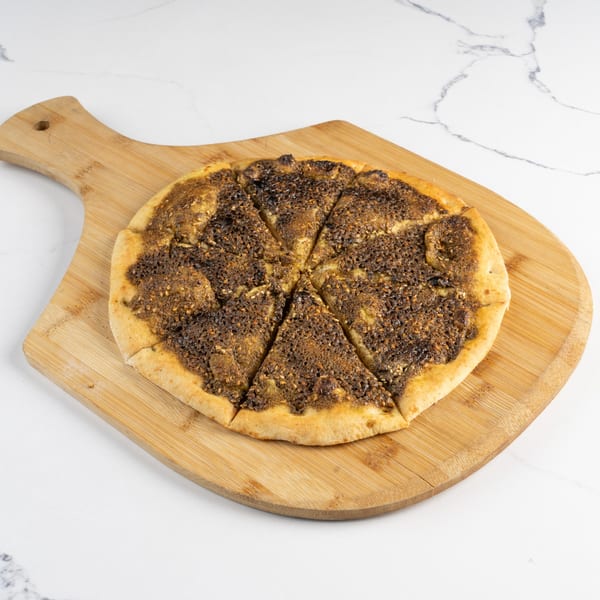 A round, sliced za'atar manakish on a wooden pizza peel against a white marble background.
