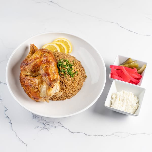 Golden brown roasted chicken with rice, lemon slices, and sides of pickles and garlic sauce.